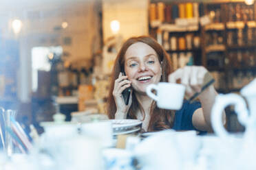 Lächelnde rothaarige Frau, die mit ihrem Smartphone telefoniert und in einem Geschäft Geschirr einkauft - KNSF09706