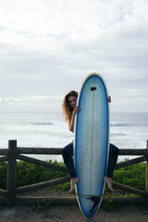 Junge Frau mit Surfbrett vor dem Meer sitzend - DCRF01755