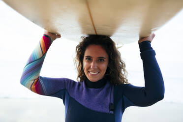 Lächelnde junge Frau mit Surfbrett auf dem Kopf am Strand - DCRF01765