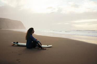 Nachdenkliche Frau mit Surfbrett am Strand sitzend - DCRF01769