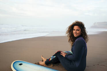 Lächelnde Frau sitzt mit Surfbrett am Strand im Urlaub - DCRF01770