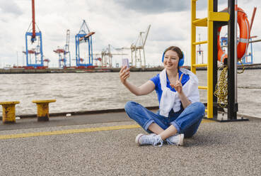Smiling teenager taking selfie on smart phone sitting on pier - IHF01604