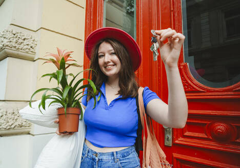 Smiling teenage girl carrying potted plant, pillow and apartment keys - IHF01608