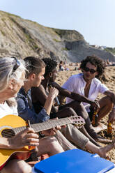 Älterer Mann mit Gitarre sitzt bei Freunden und trinkt Bier am Strand - PBTF00137