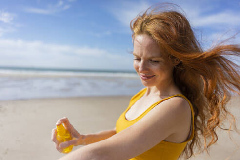 Smiling woman spraying suntan lotion on hand at beach - KNSF09708