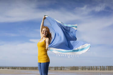 Glückliche Frau spielt mit Schal am Strand an einem sonnigen Tag - KNSF09723