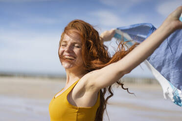 Cheerful woman with tousled hair playing with scarf on beach holiday - KNSF09724