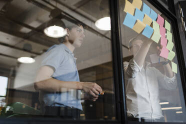 Businessman discussing with colleague sticking adhesive notes on glass wall at office - DCRF01788