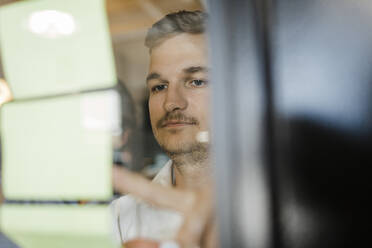 Focused businessman looking at adhesive wall on glass wall at office - DCRF01793