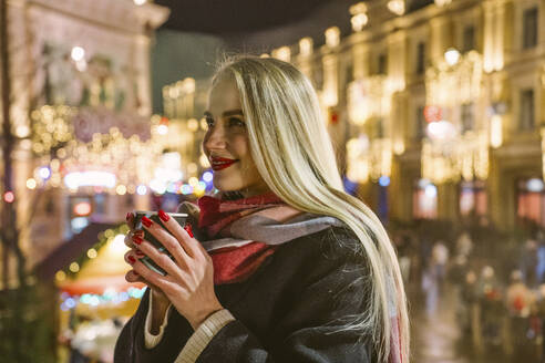 Glückliche blonde Frau mit langen Haaren hält eine Tasse Glühwein auf dem Weihnachtsmarkt - YBF00114