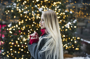 Glückliche blonde Frau vor einem Weihnachtsbaum stehend - YBF00116