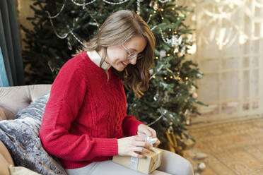 Smiling woman tying ribbon on Christmas present at home - ONAF00625