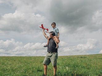 Son on father's shoulders playing with toy airplane on grassy land - VSNF01326