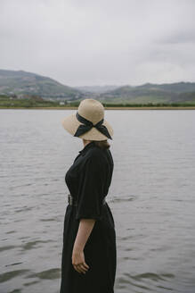 Nachdenkliche Frau in schwarzem Kleid und Strohhut am See stehend - YBF00118