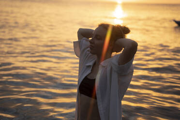 Junge Frau mit Händen hinter dem Kopf entspannt am Meer am Strand - YBF00148