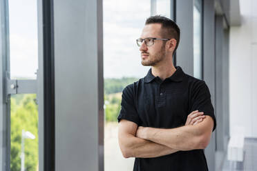 Ein nachdenklicher Geschäftsmann steht am Fenster, die Arme verschränkt, in Gedanken versunken und blickt in die Ferne - DIGF20372