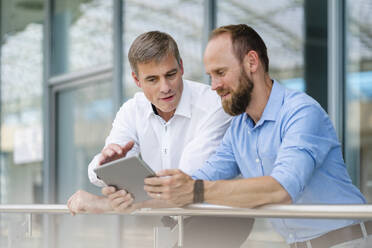 Kollegen besprechen eine Geschäftsstrategie, während sie sich auf ein Geländer eines Bürogebäudes stützen und ein digitales Tablet benutzen - DIGF20410