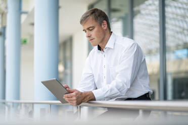 Ein Geschäftsmann lehnt sich an das Geländer eines Bürogebäudes, während er ein digitales Tablet benutzt - DIGF20411