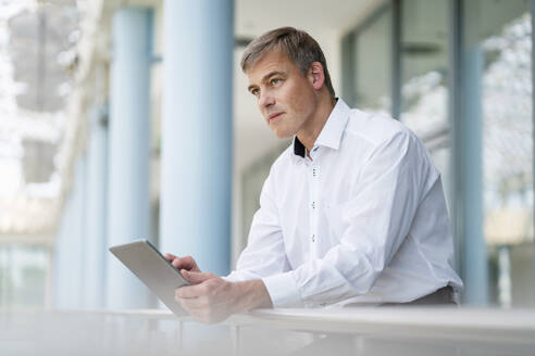 Ein ruhiger Geschäftsmann benutzt ein digitales Tablet, während er sich auf ein Geländer in einem Bürogebäude stützt - DIGF20412