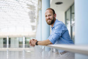 Selbstbewusster Geschäftsmann, der auf dem Balkon eines Bürogebäudes steht, sich auf das Geländer stützt und auf die Stadtlandschaft blickt - DIGF20437