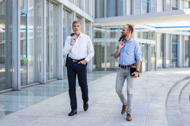 Zwei leitende Angestellte diskutieren über Geschäfte, während sie durch ein modernes Bürogebäude schlendern - DIGF20448