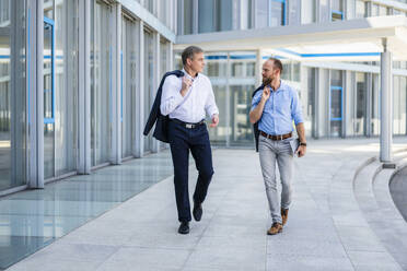 Zwei leitende Angestellte besprechen Geschäfte, während sie durch ein elegantes, modernes Bürogebäude schlendern - DIGF20450