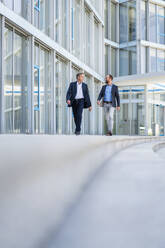 Zwei leitende Angestellte besprechen Geschäfte, während sie durch ein elegantes, modernes Bürogebäude schlendern - DIGF20456