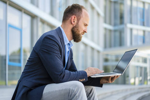 Ein konzentrierter Geschäftsmann arbeitet an seinem Laptop, während er vor einem modernen Bürogebäude sitzt - DIGF20484