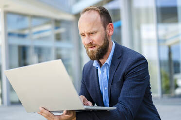 Ein konzentrierter Geschäftsmann arbeitet mit seinem Laptop, während er vor einem modernen Bürogebäude sitzt - DIGF20485