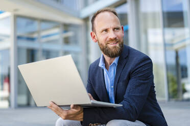 Ein konzentrierter Geschäftsmann arbeitet mit seinem Laptop, während er vor einem modernen Bürogebäude sitzt - DIGF20486