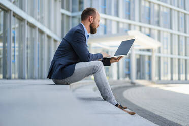 Ein Geschäftsmann arbeitet an seinem Laptop, während er vor einem modernen Bürogebäude sitzt - DIGF20487