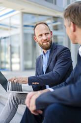 Zwei Fachleute arbeiten an einem Laptop, während sie auf Stufen sitzen und Geschäftsstrategien und -ideen diskutieren - DIGF20493