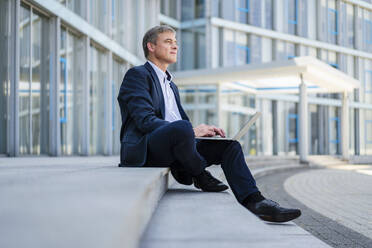 Businessman sitting in front of office building using laptop - DIGF20495