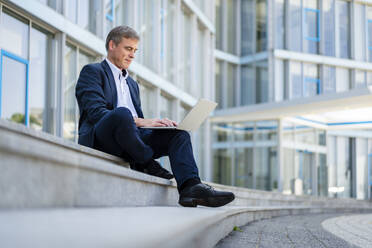 Businessman sitting in front of office building using laptop - DIGF20496