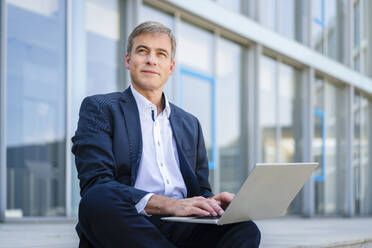 Ein konzentrierter Geschäftsmann arbeitet an seinem Laptop, während er vor einem modernen Bürogebäude sitzt - DIGF20497