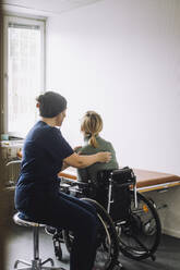 Side view of female nurse examining woman sitting on wheelchair in clinic - MASF37510