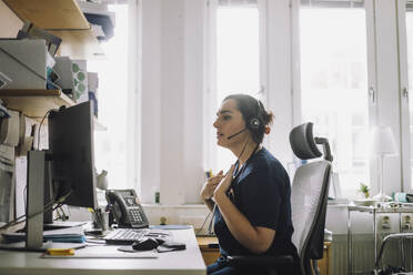 Side view of mature female nurse doing video call while sitting on chair in clinic - MASF37520