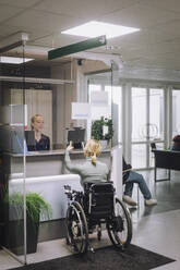 Rear view of female patient with disability doing inquiry at hospital reception - MASF37550