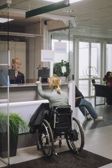 Female patient sitting on wheelchair and doing inquiry with receptionist at healthcare center - MASF37551