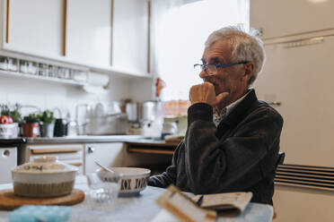 Thoughtful senior man with hand on chin sitting in kitchen at home - MASF37600