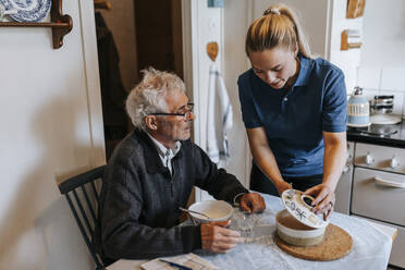 Weibliche Pflegekraft serviert Essen für einen älteren Mann, der zu Hause am Esstisch sitzt - MASF37602