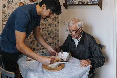 Männlicher Pfleger serviert Essen für einen älteren Mann, der zu Hause am Esstisch sitzt - MASF37603