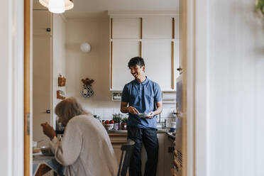 Lächelnder männlicher Pfleger im Gespräch mit einer älteren Frau beim Essen in der Küche zu Hause - MASF37605