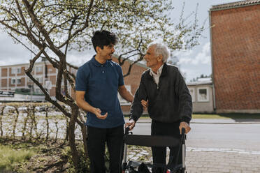 Smiling male caregiver assisting senior man with walker on footpath - MASF37621