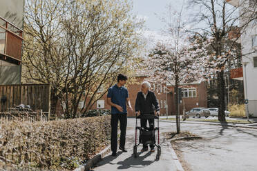 Männlicher Pflegehelfer im Gespräch mit einem älteren Mann mit Rollator auf dem Gehweg - MASF37622