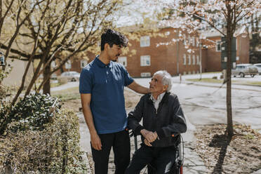 Männlicher Pfleger mit Hand auf der Schulter eines älteren Mannes, der auf einer Gehhilfe sitzt - MASF37623