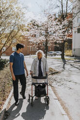 Male caregiver walking with senior woman using walker on sidewalk - MASF37624