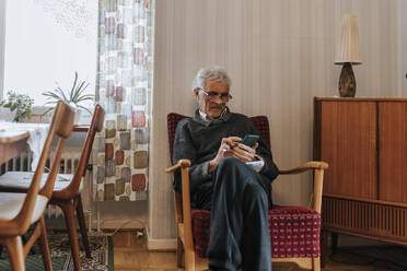 Senior man using smart phone while sitting on chair at home - MASF37639