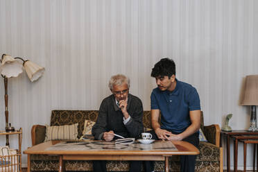 Senior man discussing with male caregiver sitting on sofa at home - MASF37641