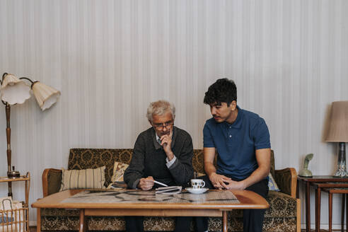 Älterer Mann diskutiert mit männlichem Betreuer, der zu Hause auf dem Sofa sitzt - MASF37641